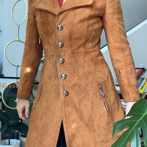 Peut inclure: Un manteau en daim marron clair avec une coupe ajust&eacute;e, un col crant&eacute;, des boutons argent&eacute;s et des poches zipp&eacute;es. Le manteau a une esth&eacute;tique vintage et un design classique, adapt&eacute; &agrave; diverses occasions.