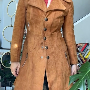 Peut inclure: Un manteau en daim marron avec une fermeture boutonn&eacute;e, des revers crant&eacute;s et deux poches zipp&eacute;es. Le manteau a une coupe ajust&eacute;e et arrive au genou. Le mod&egrave;le porte un haut &agrave; motifs rouges et blancs en dessous. Un manteau vintage &eacute;l&eacute;gant.