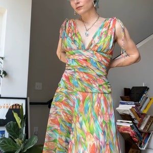 Può includere: Una donna con capelli corti blu indossa un abito senza maniche con stampa floreale colorata e scollo a V. L'abito ha un corpetto aderente e una gonna fluida.