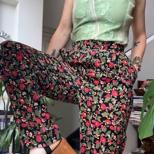 May include: A sleeveless green top with floral embroidery is paired with high-waisted black trousers featuring a red floral pattern. Brown platform sandals complete the outfit, showcasing a vintage or bohemian style.