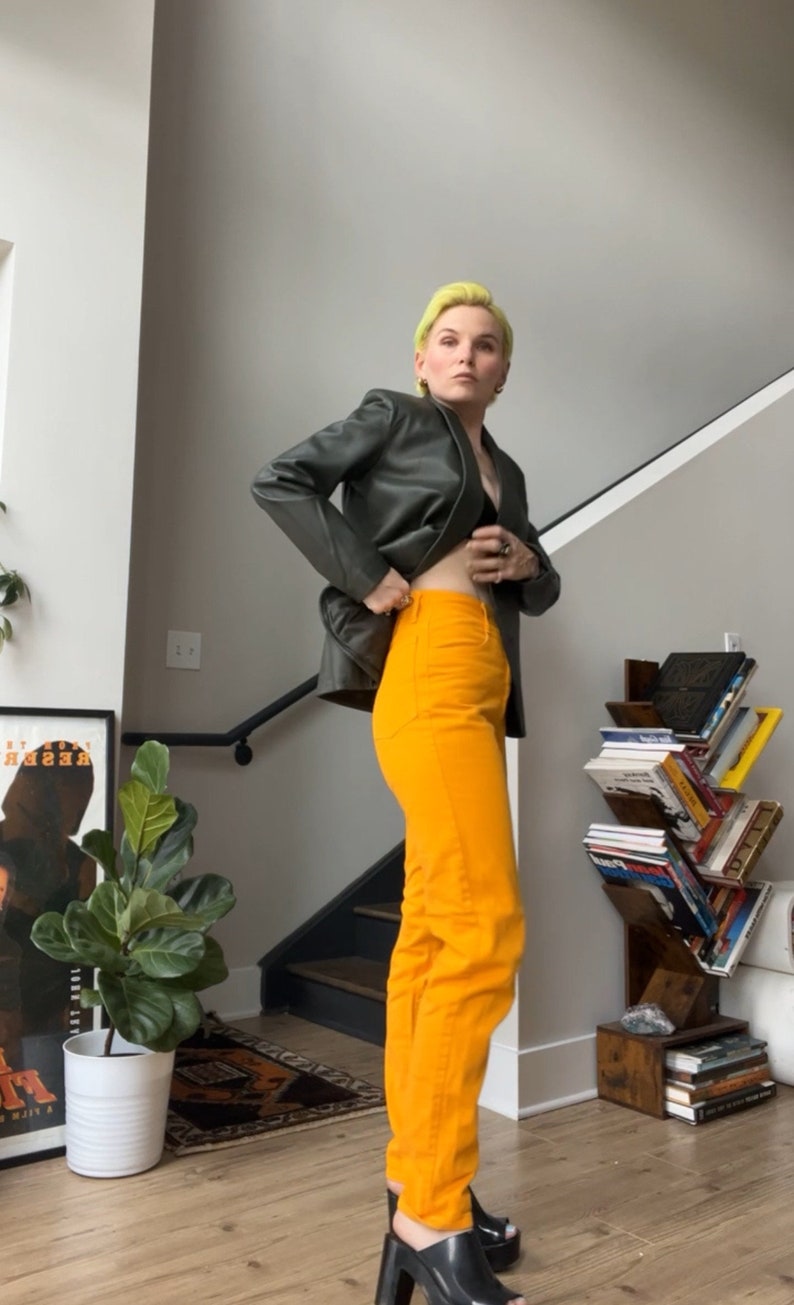 May include: A person wearing a dark green leather jacket, bright orange jeans, and black platform heels. The person has short, bright yellow hair.