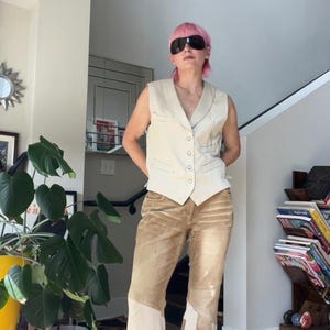 Puede incluir: Un chaleco sin mangas de color crema con cuello en V y cierre de botones frontal, combinado con pantalones anchos con un diseño de patchwork en tonos bronceado y crema. Los pantalones tienen un aspecto desgastado con paneles geométricos.