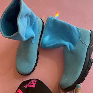 May include: A pair of light blue suede boots with black soles. The boots have a soft, folded-over cuff and a zipper detail. The boots are set against a pink background, with a pair of black Adidas track pants with pink stripes.
