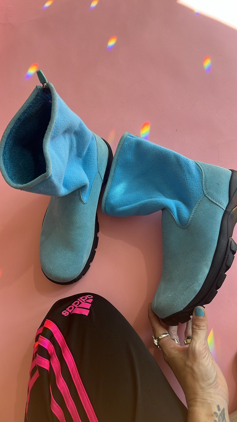 May include: A pair of light blue suede boots with black soles. The boots have a soft, folded-over cuff and a zipper detail. The boots are set against a pink background, with a pair of black Adidas track pants with pink stripes.