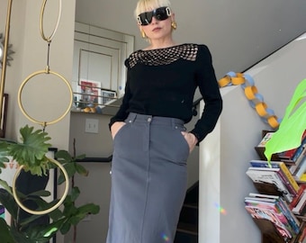 Könnte beinhalten: Eine Frau trägt ein schwarzes Langarm-Oberteil mit Netzkragen und einen grauen Maxirock. Das Outfit wird durch eine schwarze Sonnenbrille und schwarze Stiefel mit Absatz ergänzt. Goldene Ohrringe sind sichtbar. Der Hintergrund umfasst eine Treppe und Zimmerpflanzen.