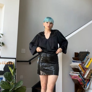 May include: A woman with blue hair is wearing a black long-sleeved blouse with a ruffled neckline and a black sequined mini skirt. She is wearing white cowboy boots with gold accents.