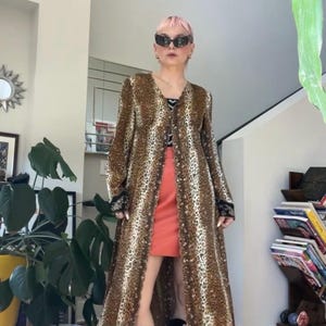 May include: A long, open-front leopard print duster coat with long sleeves and black lace trim at the cuffs, worn over a black and white patterned top, a solid coral-colored mini skirt, and black lace-up platform boots with cut-out details.