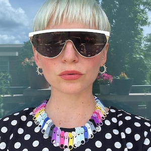 Può includere: Una persona con capelli corti e biondi chiari indossa una camicia a pois neri e bianchi. Indossa una collana fatta di cerniere colorate. La persona indossa anche grandi occhiali da sole scuri con montatura bianca.