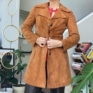 Peut inclure: Un manteau en daim marron avec une fermeture boutonn&eacute;e, deux poches zipp&eacute;es et une ceinture assortie. Le manteau est port&eacute; avec un pantalon en cuir noir et une casquette en cuir marron. L'ensemble est compl&eacute;t&eacute; par des lunettes de soleil.
