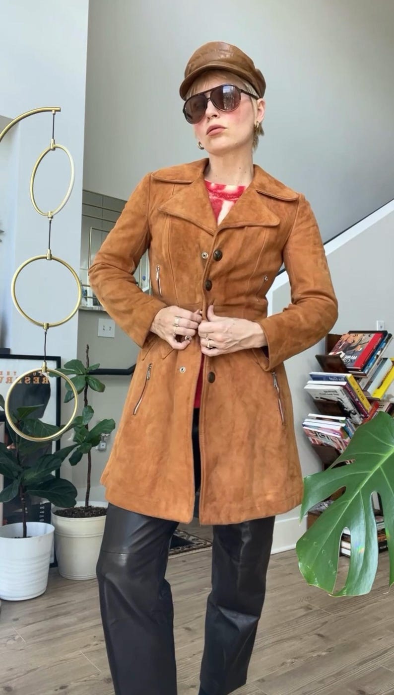 Peut inclure: Un manteau en daim marron avec une fermeture boutonn&eacute;e, deux poches zipp&eacute;es et une ceinture assortie. Le manteau est port&eacute; avec un pantalon en cuir noir et une casquette en cuir marron. L'ensemble est compl&eacute;t&eacute; par des lunettes de soleil.
