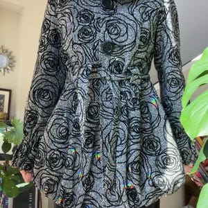 May include: A black and white coat with a rose pattern. The coat has a large collar, two large buttons, and a belt at the waist. The coat's skirt flares out from the waist. The sleeves are long and end in a ruffle.