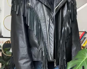 May include: Black leather jacket with fringe detailing. The jacket features a classic biker style with a notched collar, a zipper closure, and fringe accents along the sleeves and chest. The model is wearing a black top and blue jeans.
