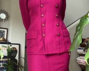 May include: A vibrant fuchsia two-piece suit. The blazer features a double-breasted design with gold-toned buttons and two front pockets. The matching skirt is a classic, tailored style. The suit is made of a textured fabric.