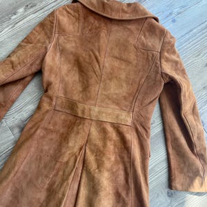 Peut inclure: Un manteau vintage en daim marron. Le manteau a un design classique avec un col crant&eacute;, des manches longues et une coupe ajust&eacute;e. Le dos a un d&eacute;tail de ceinture et une jupe pliss&eacute;e. Le manteau est de ton chaud et terreux.