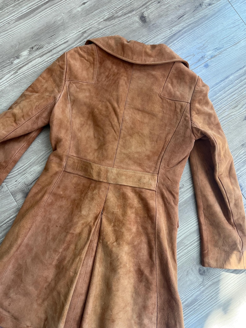 Peut inclure: Un manteau vintage en daim marron. Le manteau a un design classique avec un col crant&eacute;, des manches longues et une coupe ajust&eacute;e. Le dos a un d&eacute;tail de ceinture et une jupe pliss&eacute;e. Le manteau est de ton chaud et terreux.