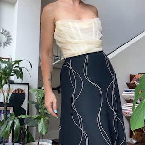 May include: A strapless cream-colored top paired with a long, flowing black skirt. The skirt features a white, wavy pattern. The outfit is a stylish ensemble, perfect for a special occasion or a night out. The model is standing in a well-lit room.