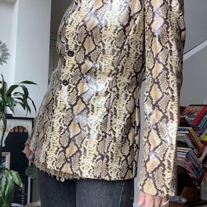 May include: A long-sleeved blazer featuring a tan and dark brown snakeskin print pattern. The garment has a high collar, a structured fit, and two dark buttons centred on the front. It is paired with dark grey trousers to complete the look.
