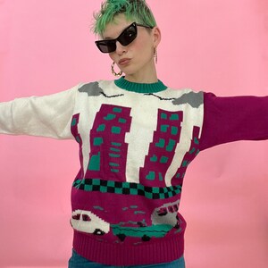 Pu&ograve; includere: Un maglione bianco, rosa e verde con un design di skyline cittadino e auto. Il maglione ha un collo rotondo e maniche lunghe.