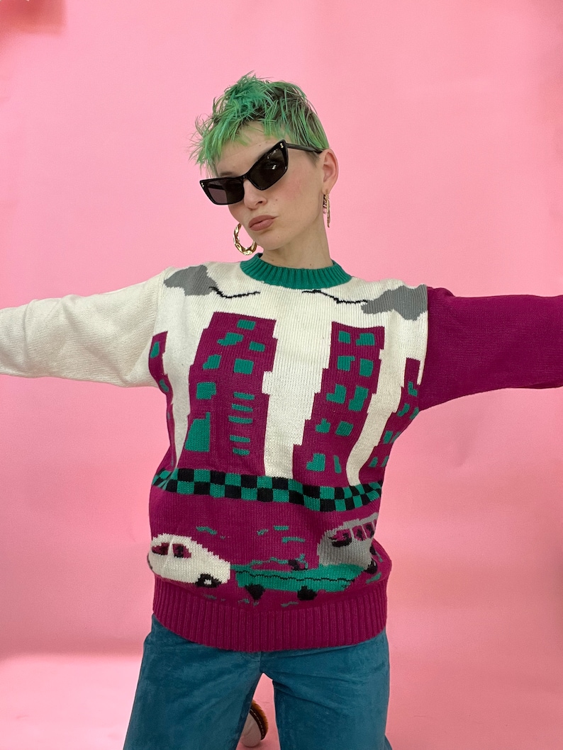 Pu&ograve; includere: Un maglione bianco, rosa e verde con un design di skyline cittadino e auto. Il maglione ha un collo rotondo e maniche lunghe.