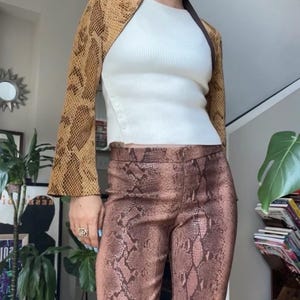 Puede incluir: Una mujer con una chaqueta y pantalones con estampado de piel de serpiente. La chaqueta es marrón y beige, y los pantalones son de un color similar. Debajo, se ve un top blanco acanalado. El conjunto se completa con gafas de sol.