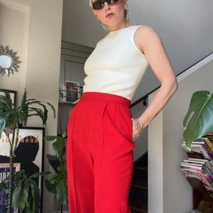 May include: A person wearing a white ribbed tank top and high-waisted, bright red trousers. The outfit is paired with dark sunglasses. The background includes indoor plants and a staircase.