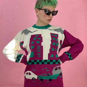 Pu&ograve; includere: Un maglione di lana bianco, rosa e verde con un design di skyline e auto. Il maglione ha un collo rotondo e maniche lunghe.