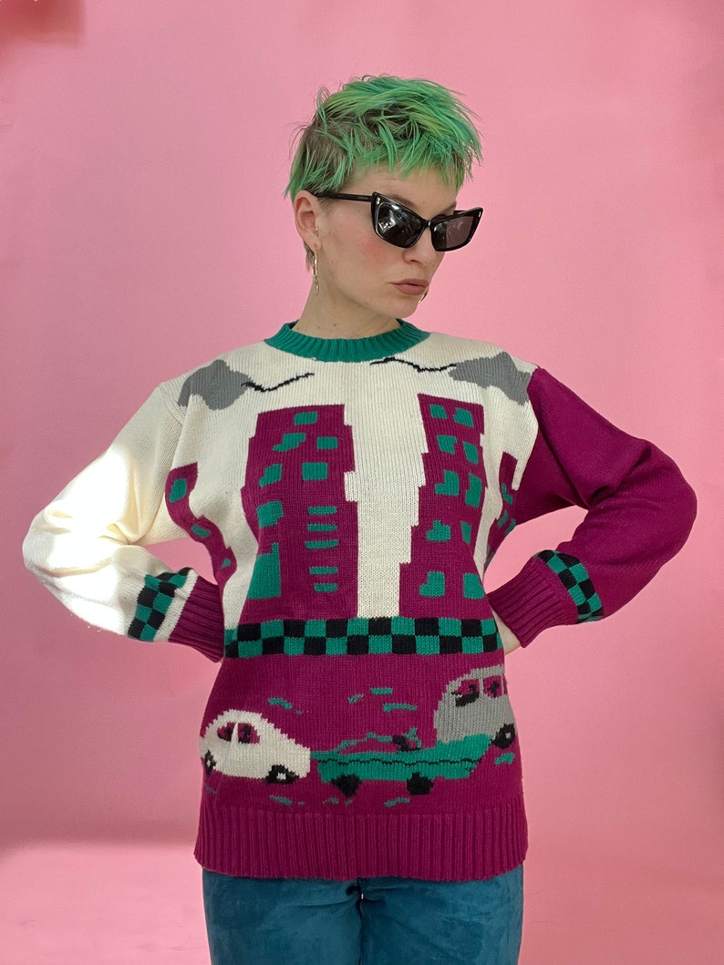 Pu&ograve; includere: Un maglione di lana bianco, rosa e verde con un design di skyline e auto. Il maglione ha un collo rotondo e maniche lunghe.