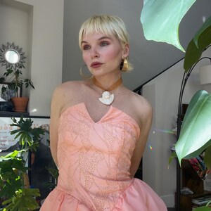 以下が含まれることがあります： ハートネックラインとレイヤードスカートが特徴の、ストラップレスのピーチ色のドレス。ボディスには、繊細なドット模様が施されています。白いペンダントが付いたゴールドトーンのネックレスが着用されています。モデルは短いブロンドの髪とゴールドのフープイヤリングをしています。