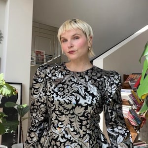 以下が含まれることがあります： 黒と銀色の花柄の長袖トップス。 トップスは丸いネックラインとパフショルダーです。 生地は光沢のあるメタリックな外観です。 この衣服はヴィンテージスタイルのブラウスです。
