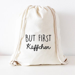 Puede incluir: Una bolsa de cordón blanco con el texto "BUT FIRST KAFFCHEN" impreso en negro.