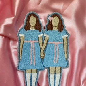 Peut inclure: Deux figures brodées en feutre de filles en robes bleues avec des rubans roses. Les filles ont les cheveux bruns et se tiennent la main.