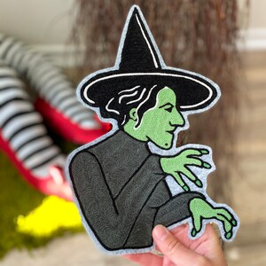 May include: An embroidered felt patch depicting a green witch with black hair and a black pointed hat.