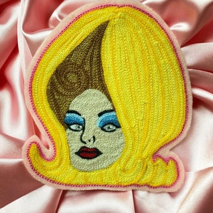 May include: Embroidered patch of a woman with blonde hair, blue eyes, and red lips. The patch is yellow, brown, and pink.