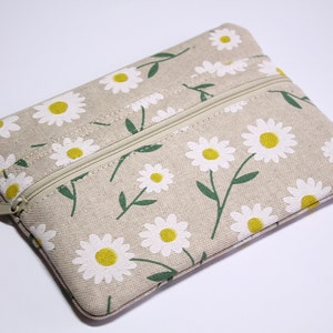 Könnte beinhalten: Eine beige Reißverschlusstasche mit einem weißen Gänseblümchen-Druck. Die Tasche hat zwei Reißverschlüsse.