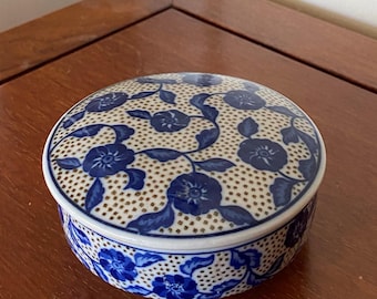 May include: A round ceramic trinket box featuring a white base with a blue floral pattern and a gold-colored dotted grid overlay. The lid is circular and fits securely over the base, which also displays the same blue floral and gold dot design.