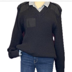 May include: Black ribbed knit sweater with a V-neck, shoulder patches, and a small square pocket. The sweater is paired with blue denim jeans. The outfit is casual and suitable for everyday wear.