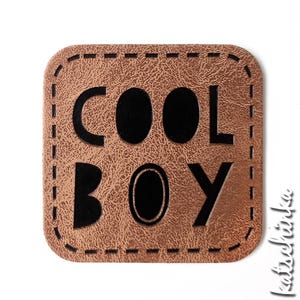 Peut inclure: Un patch en cuir marron avec une bordure cousue et un lettrage noir qui dit "COOL BOY".