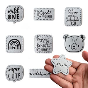 Könnte beinhalten: Neun Bügelbilder mit verschiedenen Designs und Texten in Schwarz auf grauem Hintergrund. Die Patches beinhalten einen Regenbogen, einen Bären, einen Stern, eine Sprechblase mit dem Text "wild one", ein Rechteck mit dem Text "Pfitzen Springer", ein Rechteck mit dem Text "Regen ist das Konfetti des Himmels", ein Rechteck mit dem Text "Bei Regen sind alle Schaukeln frei!", eine Sprechblase mit dem Text "super cute" und ein Rechteck mit dem Text "wirbelwind".