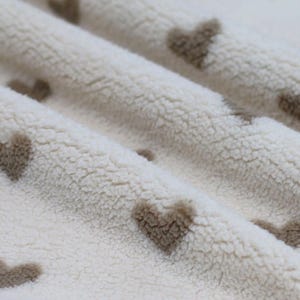 Könnte beinhalten: Nahaufnahme eines weißen Kunstpelzes mit braunem Herzmuster. Der Stoff ist weich und flauschig, und die Herzen sind gleichmäßig verteilt.