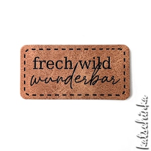 Può includere: Etichetta in pelle marrone con un bordo cucito. L'etichetta ha il testo "frech/wild wunderbar" in nero.