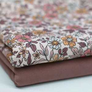 Könnte beinhalten: Zwei gefaltete Stoffstücke. Die obere Schicht zeigt ein Blumenmuster mit rosa, orangefarbenen und weißen Blumen auf hellem Hintergrund. Die untere Schicht ist einfarbig, in einem gedämpften Malvenfarbton. Der Stoff wirkt weich und eignet sich für Nähprojekte.