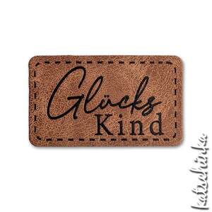 Könnte beinhalten: Ein rechteckiger Aufnäher aus braunem Kunstleder mit abgerundeten Ecken und strukturierter Oberfläche. Der Aufnäher hat schwarze Ziernähte am Rand und den schwarzen Schriftzug "Glücks Kind" in einer Kombination aus Schreib- und Serifenschrift.