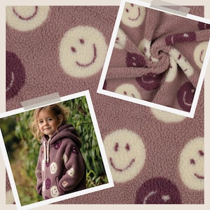 Könnte beinhalten: Nahaufnahme eines lilafarbenen Fleece-Stoffs mit einem sich wiederholenden Muster aus weißen und dunkelvioletten Smiley-Gesichtern. Das Bild zeigt auch ein Kind, das einen passenden Kapuzenpullover mit dem gleichen Muster trägt.