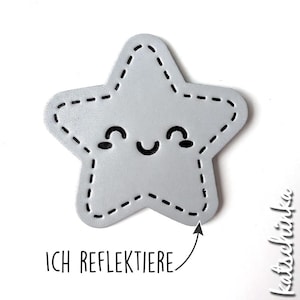 Könnte beinhalten: Ein silberner Stern mit einem gestickten Umriss und einem lächelnden Gesicht. Der Text "ICH REFLEKTIERE" befindet sich unter dem Stern.