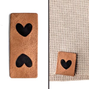 Puede incluir: Dos parches de cuero marrón con recortes de corazón negro. Un parche tiene dos corazones, el otro tiene uno. Los parches se muestran junto a una tela de punto gofrado beige.