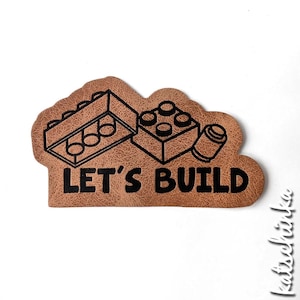 Könnte beinhalten: Brauner Leder-Patch mit schwarzem Umriss von zwei Lego-Steinen und dem Text "LET'S BUILD".