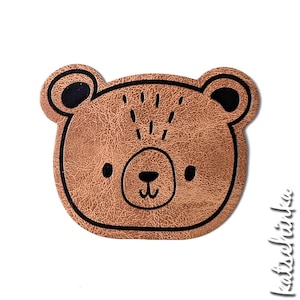 Pode incluir: Um rosto de urso de feltro marrom com olhos, nariz e boca pretos. O rosto de urso é um design simples e bonito.
