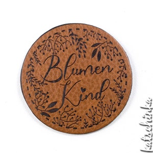 Könnte beinhalten: Ein brauner Lederkreis mit einem schwarzen floralen Design und dem Text "Blumen Kind" mit einem kleinen Herzen in der Mitte.