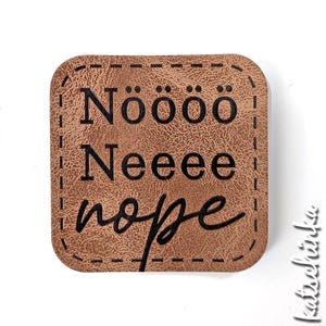 Könnte beinhalten: Ein quadratischer, hellbrauner, texturierter Kunstlederaufnäher mit abgerundeten Ecken und schwarzen Ziernähten am Rand. Der Text "Nöööö", "Neeee" und "nope" ist in Schwarz aufgedruckt. Das Wort "nope" ist in Schreibschrift.