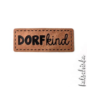 Könnte beinhalten: Ein braunes Leder-Patch mit einem gestickten Rand und dem Text "DORFKIND" in schwarzen Buchstaben.